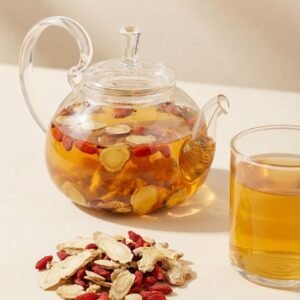 A clear glass teapot filled with brewing CozySlip Ginseng Ginger Goji tea, showing the golden amber liquid and floating ingredients, paired with a pile of dry herbs on the table.