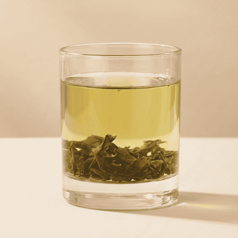 A clear glass cup filled with CozySlip Mint Green Tea, showcasing the vibrant green tea leaves settled at the bottom and the light, refreshing yellow-green liquor, set against a clean, minimalist background.