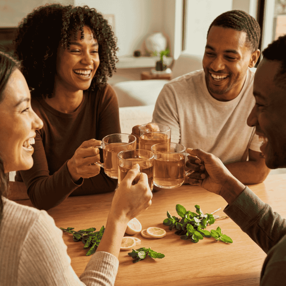A diverse group of happy friends laughing and toasting with clear glass mugs of CozySlip Mint Green Tea around a wooden table, garnished with fresh mint sprigs and lemon slices, capturing a lively afternoon social gathering.