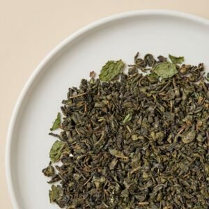 A high-resolution close-up of CozySlip Mint Green Tea loose leaf blend on a white plate, clearly showing the texture of twisted green tea leaves mixed with dried mint leaf fragments, highlighting the natural ingredients.