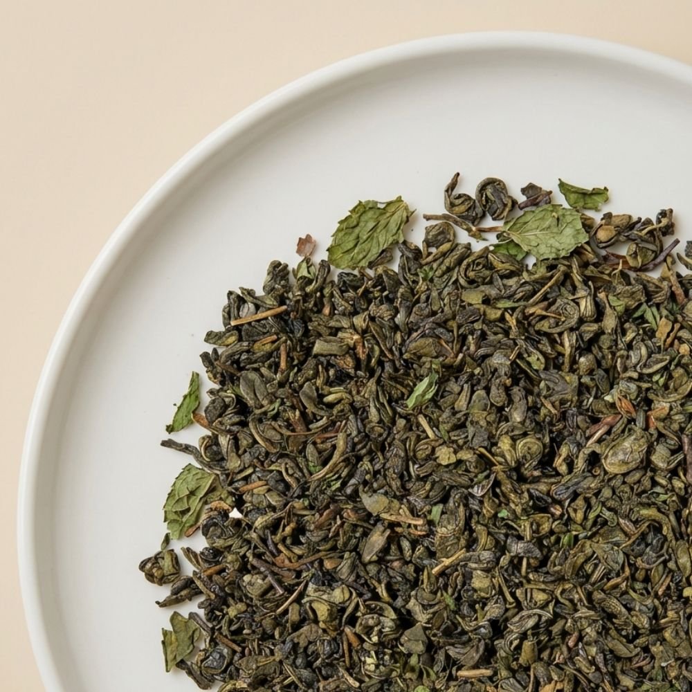 A high-resolution close-up of CozySlip Mint Green Tea loose leaf blend on a white plate, clearly showing the texture of twisted green tea leaves mixed with dried mint leaf fragments, highlighting the natural ingredients.