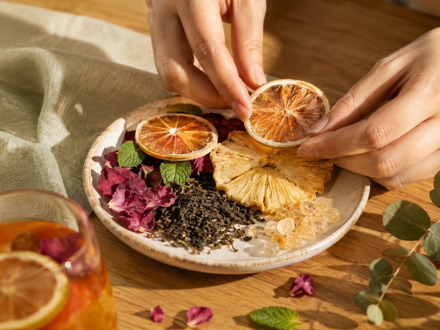 Hands arranging dried citrus, pineapple, rose, mint, tea and rock sugar for a Cozyslip fruit tea composition